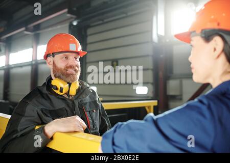 Cheerful handsome wormer in hardhat standing at railing and chatting with colleague during break at factory Stock Photo