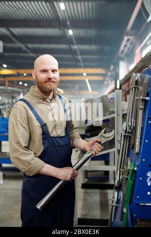 Portrait of bald bearded man holding big metal wrench and standing in industrial shop Stock Photo