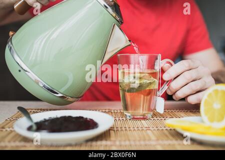 Close up focus on male hand holding kettle and pouring boiled water into glass cup - preparing hot drink for breakfast - tea or coffee Stock Photo
