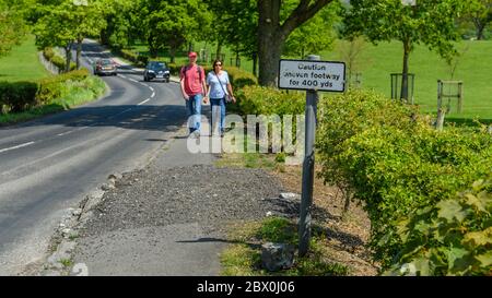 2 people walking by country lane, approaching 'Caution uneven footway' warning sign & broken damaged pavement & kerb - North Yorkshire, England, UK. Stock Photo