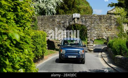 Car on scenic lane driving & low narrow stone archway (3 arches, headroom warning sign 10' 9'') - B6160, Bolton Abbey village, Yorkshire, England, UK. Stock Photo