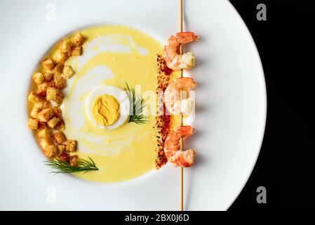Warm cream soup with cheese, egg, fresh green dill, shrimp and spices, fried croutons, in a white round plate on a black background Stock Photo