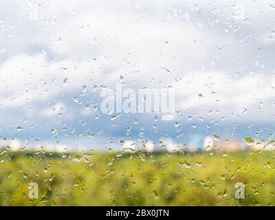 view of blurred green city park, blue cloudy sky and urban houses on horizon on sunny spring day through dirty glass of home window with raindrops (fo Stock Photo