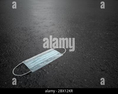 Low angle shot of white medical face protection mask for mouth and nose lying on street paved with dark grey wet tarmac on rainy day Stock Photo
