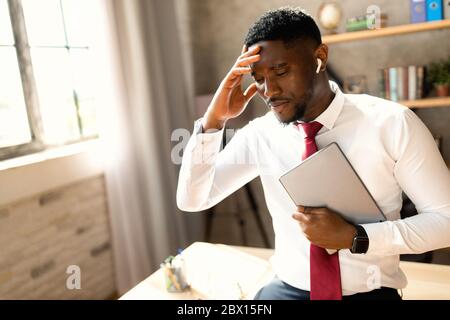 Tired young black man dressed in shirt sitting at his workplace. Tired business black man working in office dressed in white shirt, touching head with Stock Photo