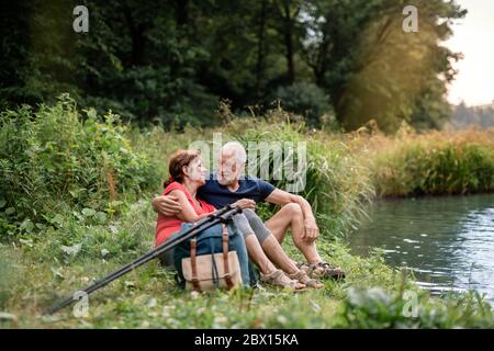 Senior tourist couple with backpacks on a walk in nature, sitting by lake.