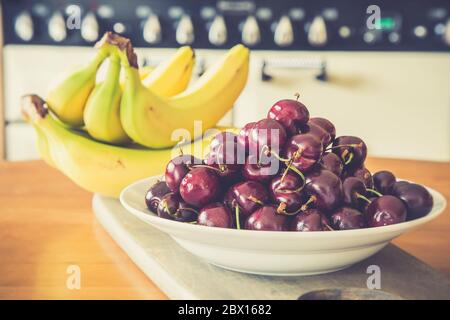 Bowl of red cherries & bunch of bananas inside on wooden UK kitchen table. Fresh fruit. Life is just a bowl of cherries. Stock Photo