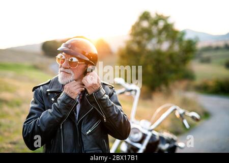 A senior man traveller with motorbike in countryside, putting on helmet.