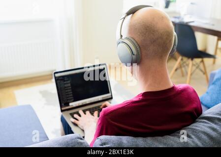 Man working from home on sofa in living room. Using laptop and headphones. Coding, programming Stock Photo