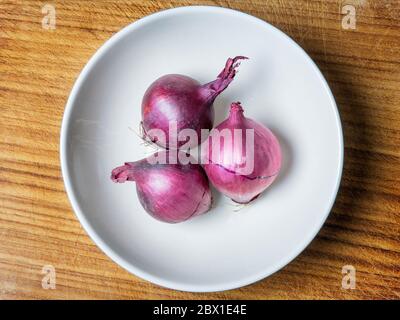 Three small red onions in a bowl on a wooden chopping board Stock Photo