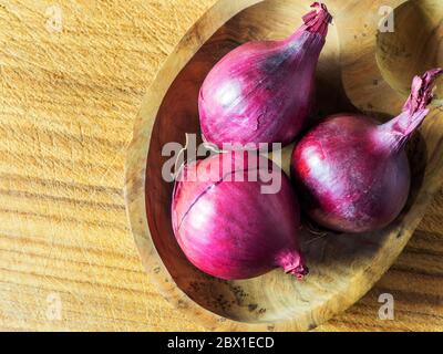 Three small red onions in a wooden dish on a wooden chopping board with copy space Stock Photo