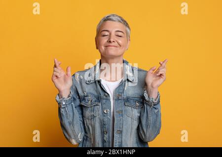 Making cherished wish. Optimistic senior lady crossing fingers with hope with closed eyes, light background with copy space Stock Photo