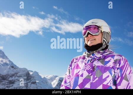 Close portrait of happy teen girl smiling wearing cute winter ski outfit glasses , helmet stand over sky and mountain peaks Stock Photo