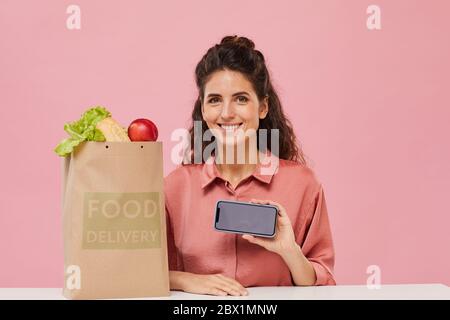 Portrait of young woman buying food online she sitting at the table and showing the screen of her mobile phone to the camera Stock Photo