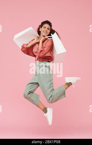 Portrait of happy shopaholic enjoying her shopping she jumping up with shopping bags against the pink background Stock Photo