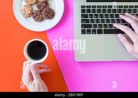 Male hands holding a cup of coffee and using laptop, cookies on colorful background. Stock Photo