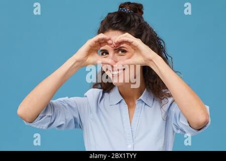 Portrait of happy young woman with curly hair making heart shape from her hands against the blue background Stock Photo