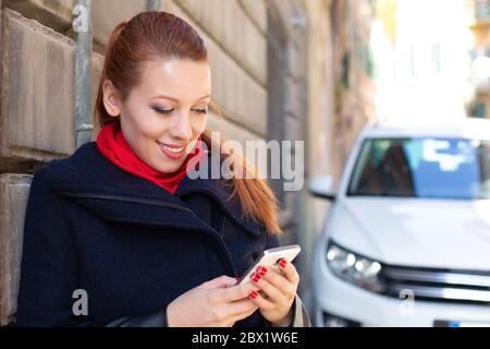 Beautiful young woman using her mobile phone, texting sending a text message leaning on a concrete wall on an Italian city street Stock Photo