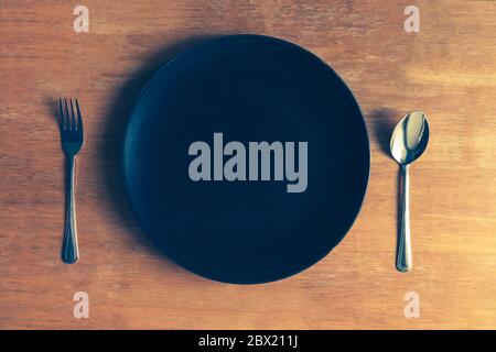 Top view of empty black plate with spoon and fork on wooden table Stock Photo