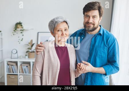 Portrait of mature bearded man caring for senior woman he embracing her and holding her hand they standing in the living room and smiling Stock Photo