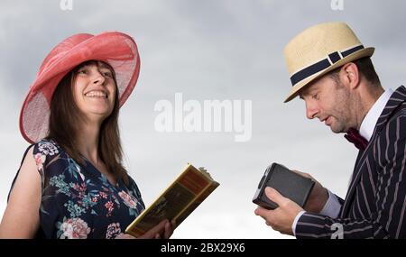 Close up UKhappy couple in vintage 1940s costume outdoors in summer sunshine. Man is photographing wife using vintage Box Brownie camera, sky backdrop. Stock Photo