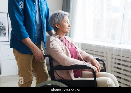 Disabled senior woman sitting in wheelchair and looking through the window with caregiver standing nearby Stock Photo