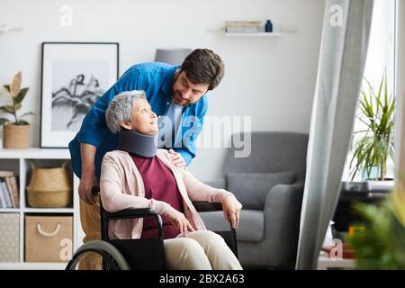 Senior woman with bandage on her neck sitting in wheelchair and talking to the man in the room Stock Photo