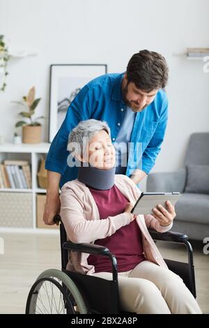 Disabled senior woman in bandage sitting in wheelchair and showing something on tablet pc to the man Stock Photo