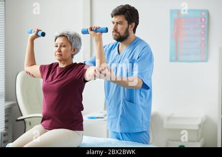 Senior woman lifting dumbbells and male doctor assisting her during rehabilitation Stock Photo