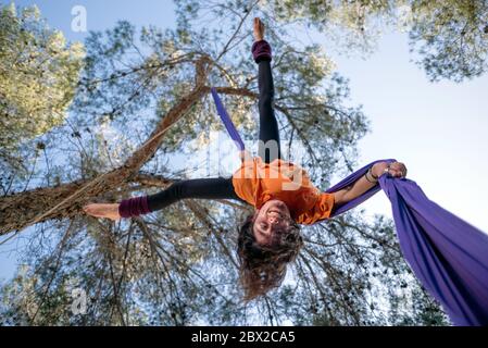 Young girl acrobat. Practicing aerial silks. Strong woman doing circus stunts with clothes in the forest. Straddle fall position. Stock Photo