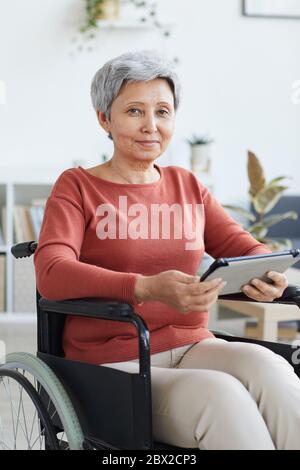 Portrait of disabled senior woman sitting in wheelchair and looking at camera while using tablet pc at home Stock Photo