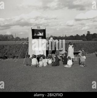 1950s, historical, a small group of children sitting on the grass at a seaside park watching a traditional Punch & Judy show, England, UK. Punch and Judy is a comedy puppet show in a booth featuring two main characters, Mr Punch and his wife Judy and other assorted characters. An historic entertainment form, it has its roots in the 16th-century Italian 'Commedia dell'arte'. Traditionally set up at or near the seaside to entertain a family audience who are on holiday, the characters are operated by a single puppeteer inside the booth. Stock Photo