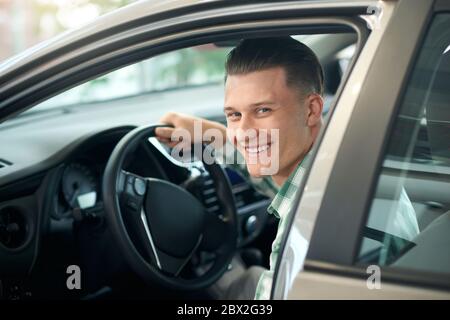 Portrait of handsome happy smiling man sitting in new car in dealership and looking at camera. Young customer feeling happy, trying sitting in comfortable vehicle before purchase, holding wheel. Stock Photo