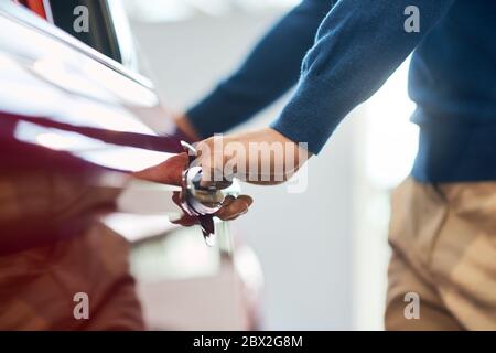 Hand of male unrecognizable businessman opening and holding red car door. Close up of young caucasian male driver in casual outfit opening door of automobile in car dealership. Stock Photo