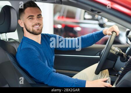 Portrait of handsome happy smiling man sitting in new car in dealership and looking at camera. Young customer feeling happy, trying sitting in comfortable vehicle before purchase, holding wheel. Stock Photo