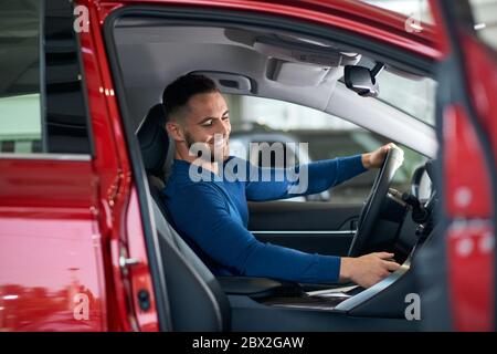 Side view of handsome happy smiling man sitting in new car in dealership and using gearbox. Young customer feeling happy, trying sitting in comfortable vehicle before purchase, holding wheel. Stock Photo