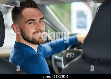 Portrait of handsome happy smiling man sitting in new car in dealership and looking at camera. Young customer feeling happy, trying sitting in comfortable vehicle before purchase, holding wheel. Stock Photo