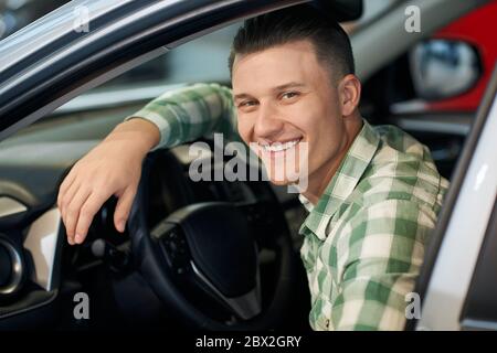 Handsome man sitting in new car in dealership, sincerely smiling with teeth and looking at camera. Young customer feeling happy, trying sitting in vehicle before purchase, leaning on wheel. Stock Photo