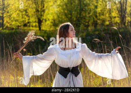 Happy medieval young woman in historical female costume at nature. Fantasy girl in long dress or gown with at field in forest Stock Photo