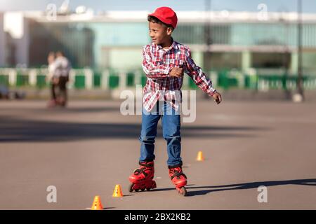 Cheerful little african american boy on rollers skates in summer park at asphalt road between training cones . Roller kid enjoying speed and smiles. Stock Photo