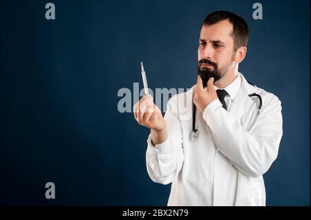 Portrait of male doctor with stethoscope in medical uniform is looking at the thermometer posing on a blue isolated background. Stock Photo