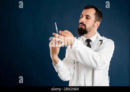 Portrait of male doctor with stethoscope in medical uniform with a syringe in hand posing on a blue isolated background. Stock Photo