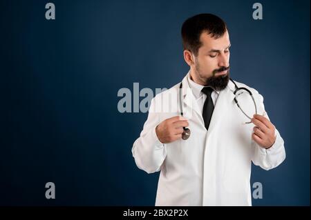 Portrait of male doctor with stethoscope in medical uniform arranges his stethoscope posing on a blue isolated background. Stock Photo