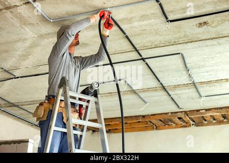 Worker electrician standing on aluminium ladder installing electrical cables laid in protective corrugation installed on the ceiling and wall in a roo Stock Photo