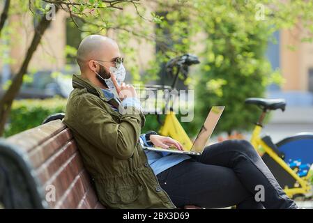 A bald man in a medical face mask to avoid the spread coronavirus works remotely on a laptop in the park. A guy wears a pilot sunglasses sits on a ben Stock Photo