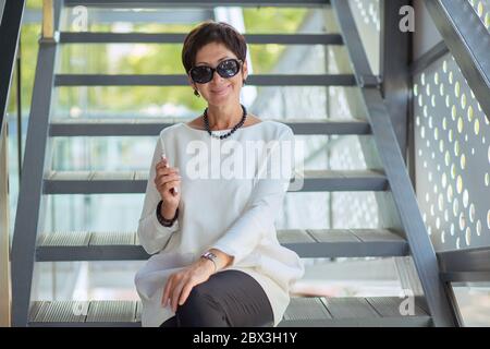 Cheerful middle-aged stylish woman in black sunglasses sitting on stairs on street relaxing looking at you camera and smiling happily wearing white bl Stock Photo