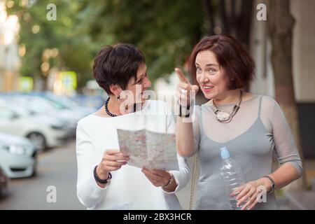 Stylish middle-aged woman with bottle of water and map standing on street looking for direction pointing to the side happy and excited while traveling Stock Photo