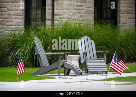 Two grey Adirondack chairs sitting on a small patio with table between them in front of tall decorative grass and windows of rock house - American fla Stock Photo