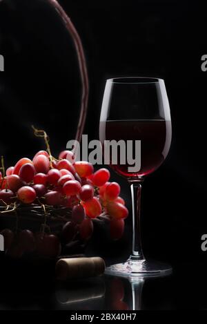 A cup of wine and a basket of grapes on the side, on a shiny surface, and in a black background. Low key light photography Stock Photo