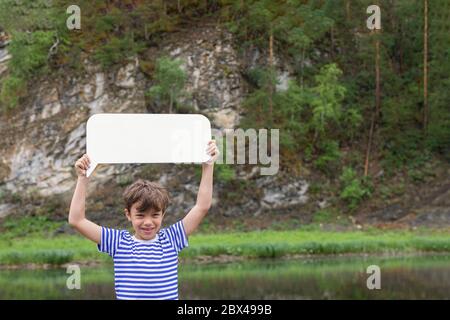 Young smiling boy standing on bank of river keeping blank white board in hands above his head, rocky cliffs on background. Summer is best time. Stock Photo